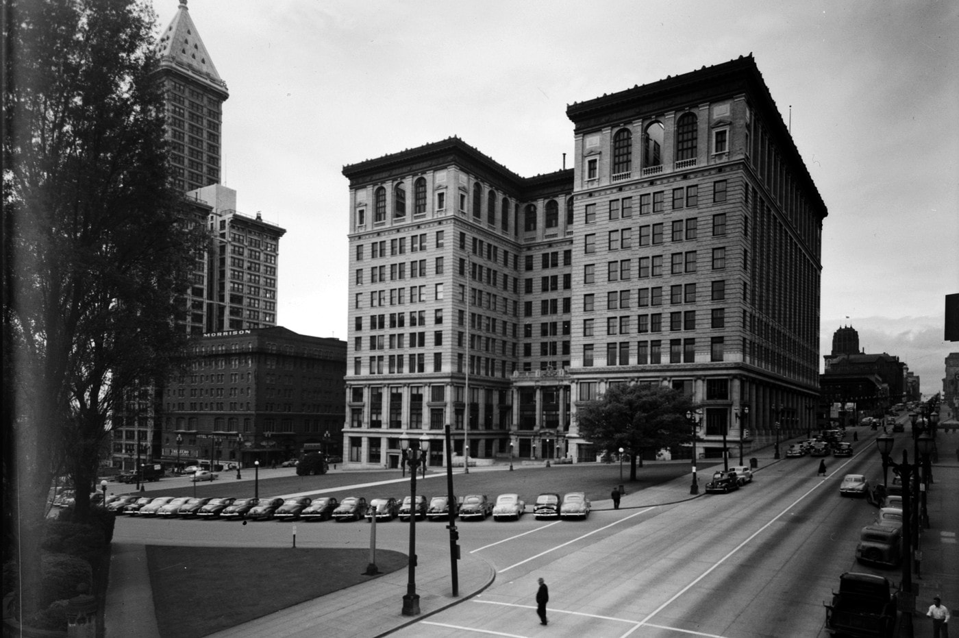 A tall stone courthouse on a city block in 1949, seen from across a park, with cars parked along the street.